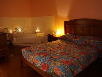 Room with pink walls and full size bed with floral bed spread. Candles line the bathtub in the corner.  
