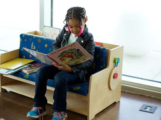 Young girl with dark braids reads a book on a bench by a window.